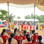 Children in bright traditional dresses perform a dance on a dirt field as an audience watches from red and brown chairs under a tented canopy